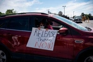 Woman in car with masks holds sign reading "Release them NOW"