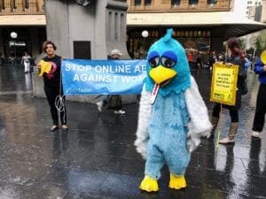 Person in Twitter costume stands with protesters in the background holding signs