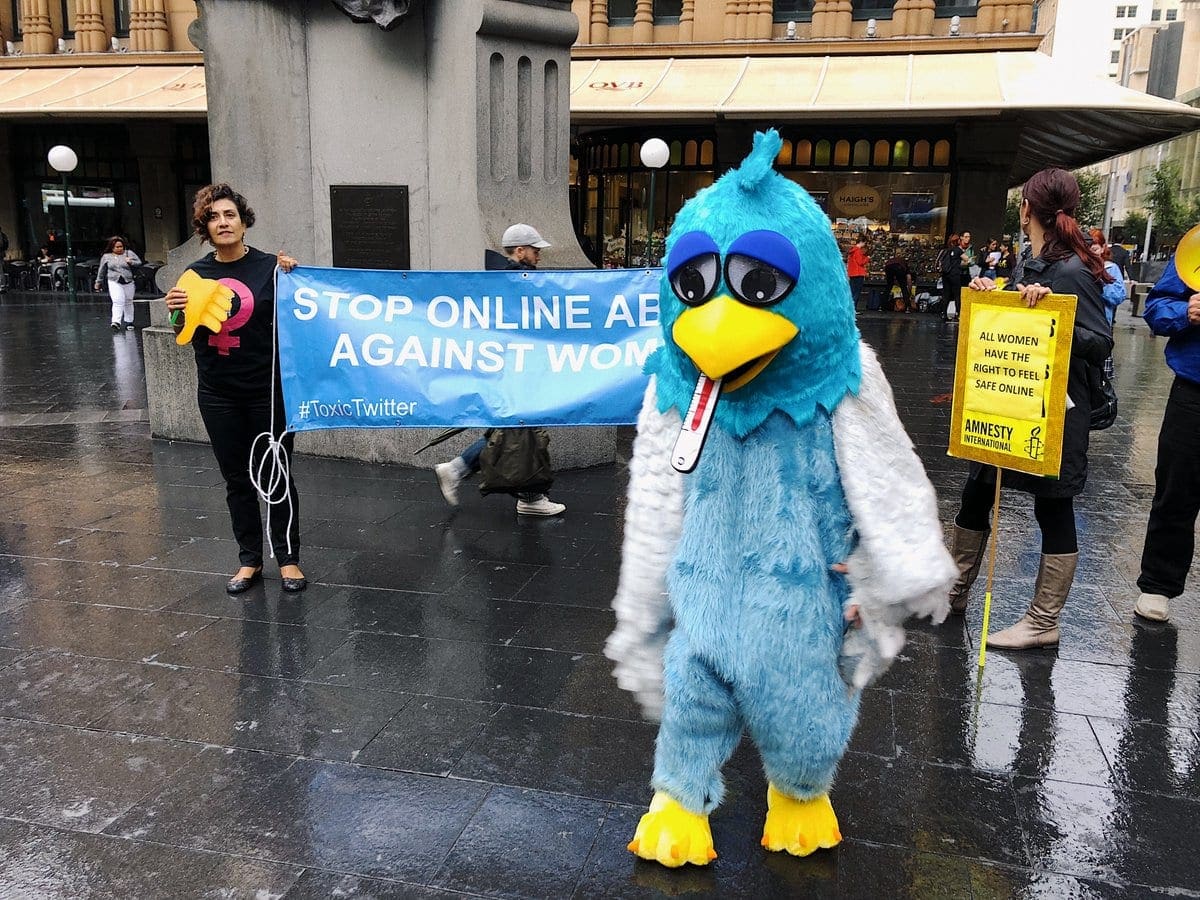 Person in Twitter costume stands with protesters in the background holding signs