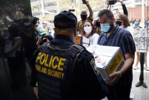 Activists wearing masks stand in front of official with uniform labeled Homeland Security