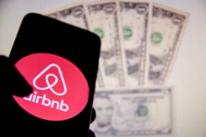 Airbnb logo is displayed on a smartphone screen with dollars in the back