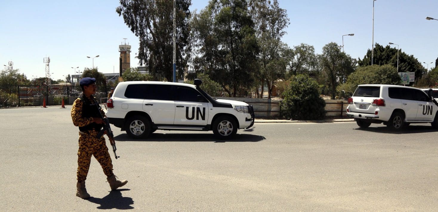 A soldier carrying an AK rifle and wearing a camoflauge uniform and a blue beret stands guard outside an airport facility, with two white UN Toyota SUVs behind him. The sky is dark blue and there are some trees behind the cars.