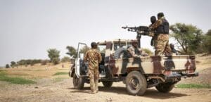 Soldiers in campflauge uniforms stand in and around a machine-gun mounted pick-up truck, also painted camoflauge, in a dry landscape with scrubby trees under a hazy blue sky. In the distance, a group of men dig pits in the sand.