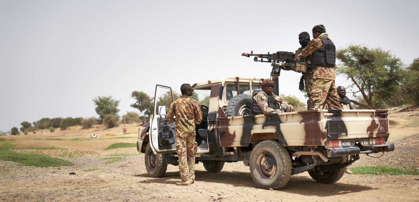 Soldiers in campflauge uniforms stand in and around a machine-gun mounted pick-up truck, also painted camoflauge, in a dry landscape with scrubby trees under a hazy blue sky. In the distance, a group of men dig pits in the sand.