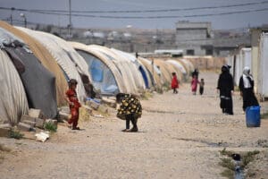 Women and children walk down a dirt path between a row of domed tents covered in plastic and canvas that stretches far into the distance under a midday sun.