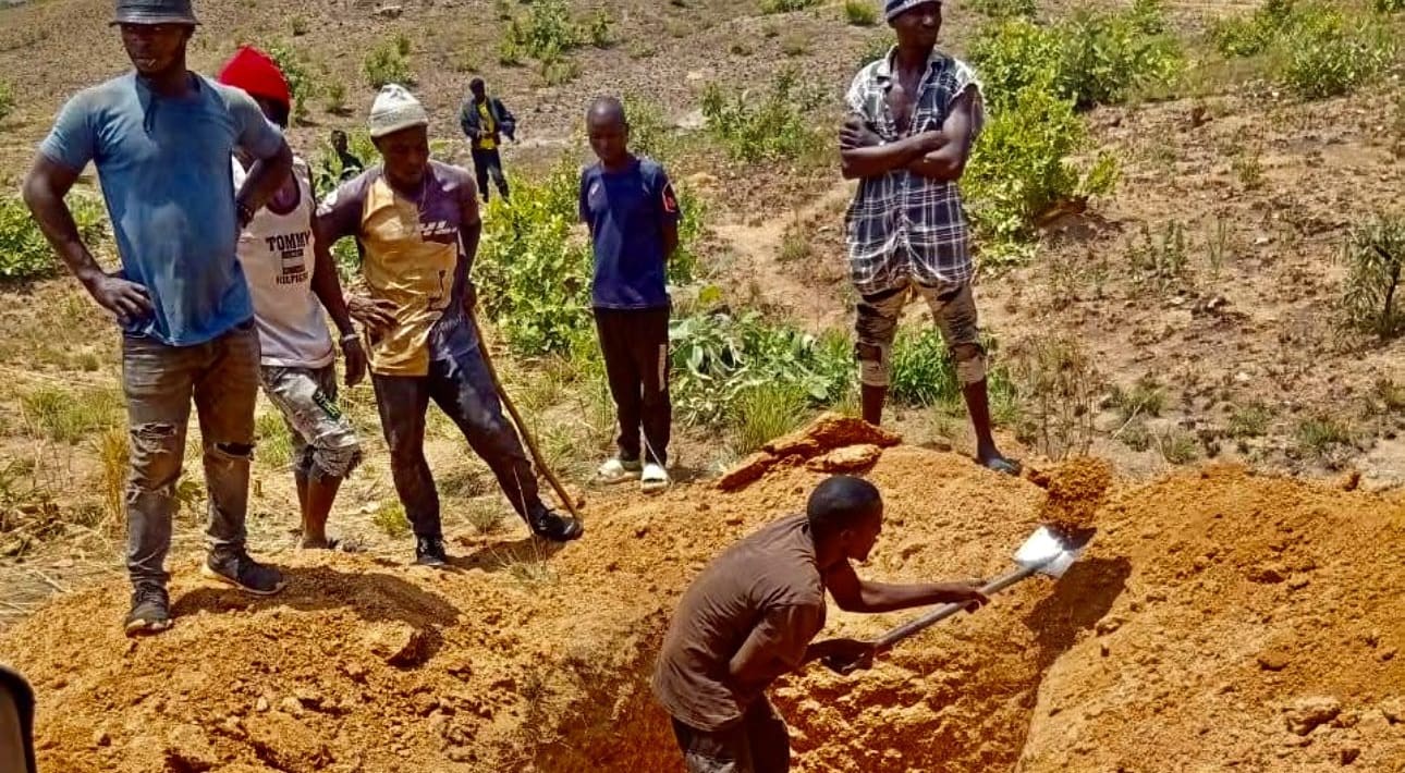 A man shovels dirt to dig a grave in dry red soil while other men watch, standing solemnly at the edge of the pit with arms crossed or hands at their hips. The shadows are harsh but small, indicating the digging takes place in the early afternoon heat.