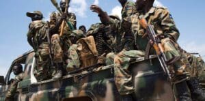 Soldiers wearing camoflauge and draped in belts of bullets sit in the back of a camo-painted picked holding AK rifles and handing rocket propelled grenades to each other.