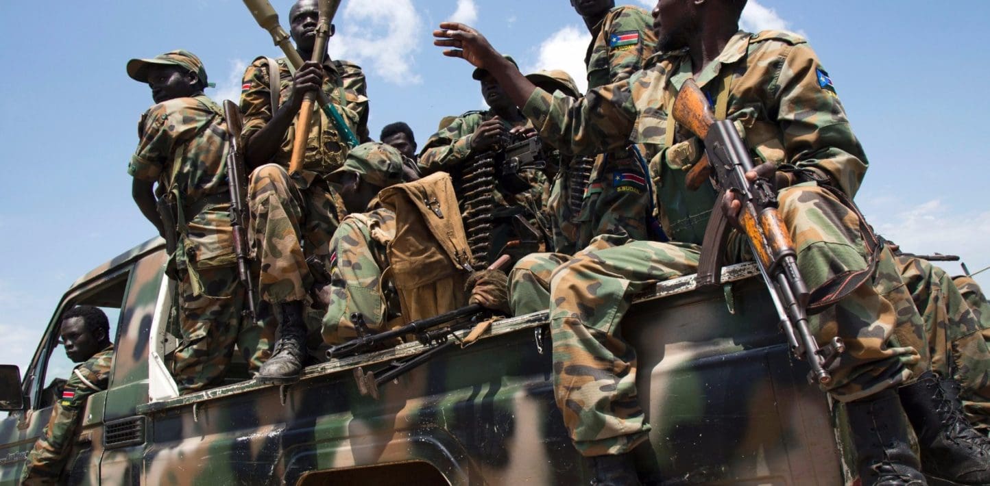 Soldiers wearing camoflauge and draped in belts of bullets sit in the back of a camo-painted picked holding AK rifles and handing rocket propelled grenades to each other.