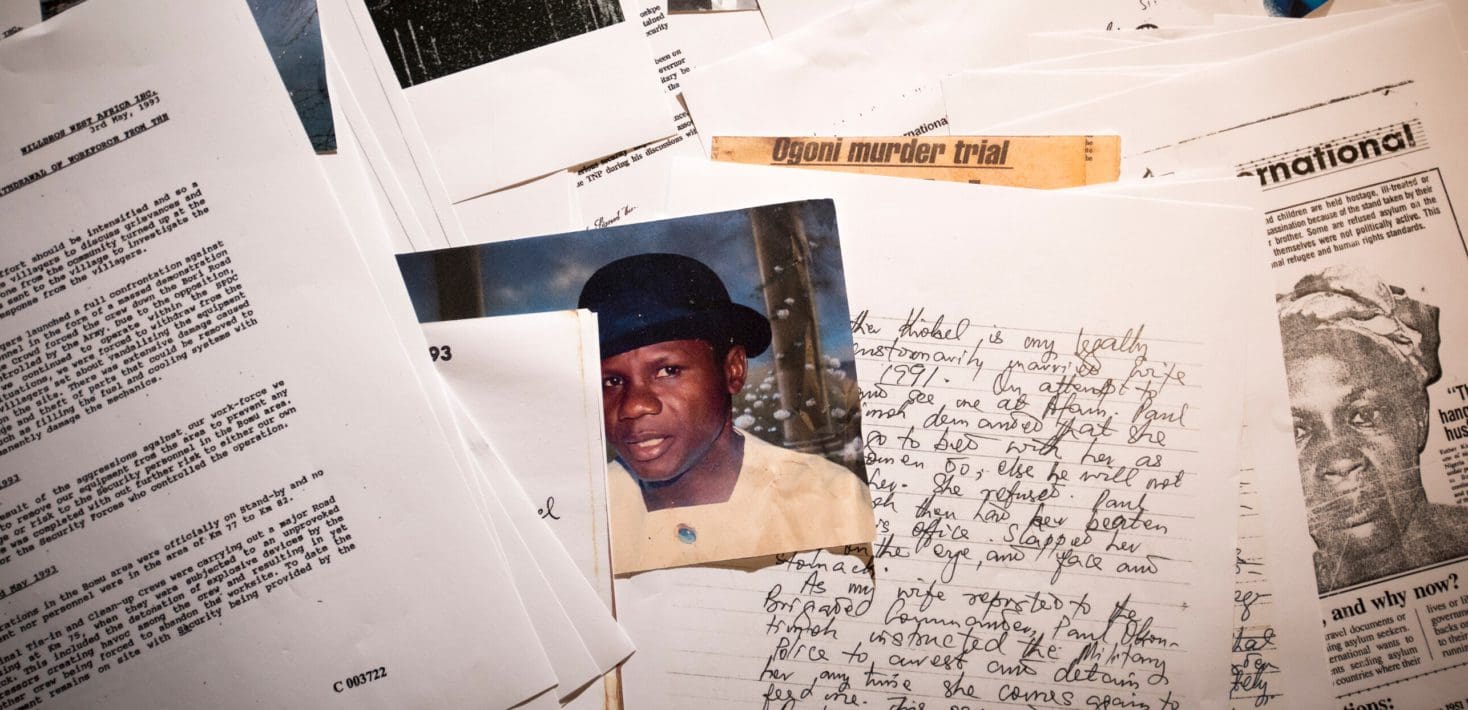 A pile of typed documents and hand written letters related to the case of the Ogoni nine, with a photo of Ken Saro-Wiwa wearing a yellow shirt and a black bowler hat.