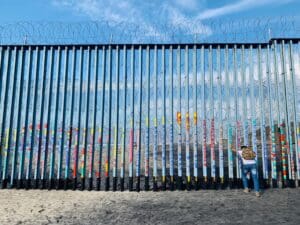 A man pushes against the tall iron bars that form the border wall between the US and Mexico at Tijuana. The ground is sandy, and there is blue sky and clouds on the US side seen through the bars as well as curled barbed wire on the US side. The bars on the Mexican side have been painted with colorful words and pictures.
