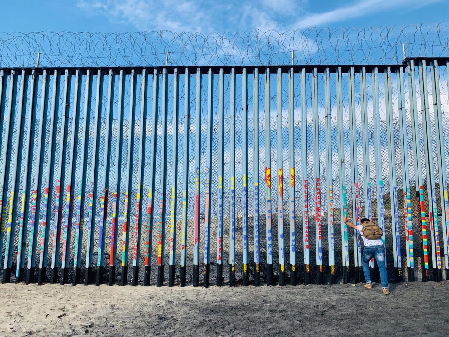 A man pushes against the tall iron bars that form the border wall between the US and Mexico at Tijuana. The ground is sandy, and there is blue sky and clouds on the US side seen through the bars as well as curled barbed wire on the US side. The bars on the Mexican side have been painted with colorful words and pictures.