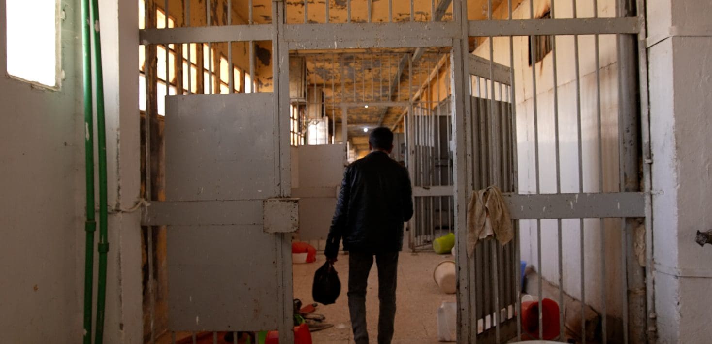 A person walks through open doors inside a hallway in a prison with bars over windows and along the cells, and buckets and shoes on the ground. The paint is peeling on the roof. The person has their back to us, and is wearing all black and carrying a small bag in their left hand.