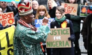 King Okabi of the Ogale community, wearing a colorful tunic and a bright red crown, stands with his fist raised before a crowd of protesters holding signs reading