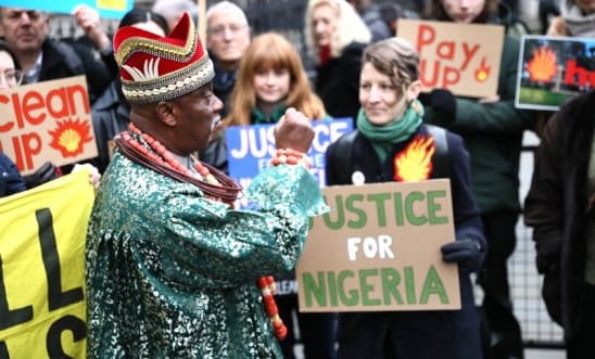 King Okabi of the Ogale community, wearing a colorful tunic and a bright red crown, stands with his fist raised before a crowd of protesters holding signs reading 