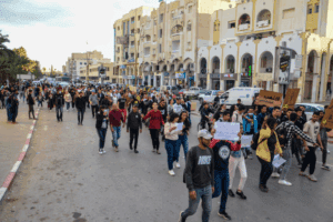 A group of young people carrying handmade cardboard signs walks down an otherwise deserted street in the late afternoon along a row of buildings with shopfronts.