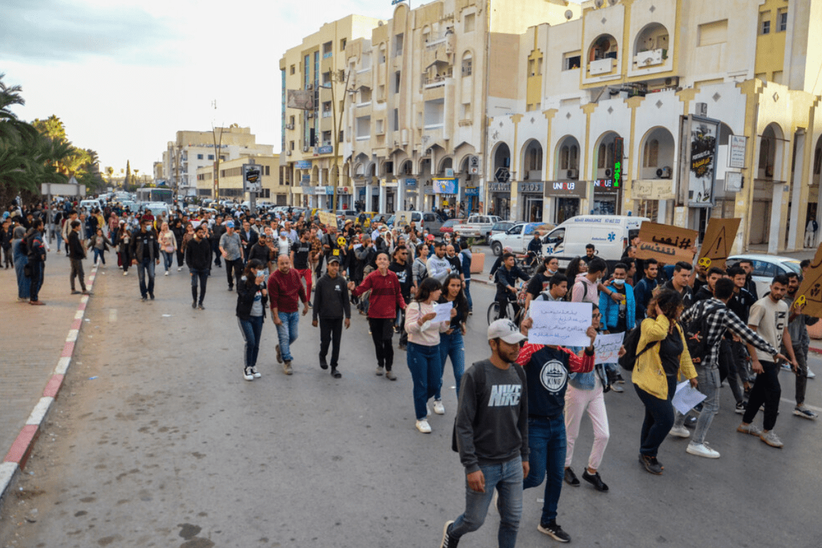 A group of young people carrying handmade cardboard signs walks down an otherwise deserted street in the late afternoon along a row of buildings with shopfronts.