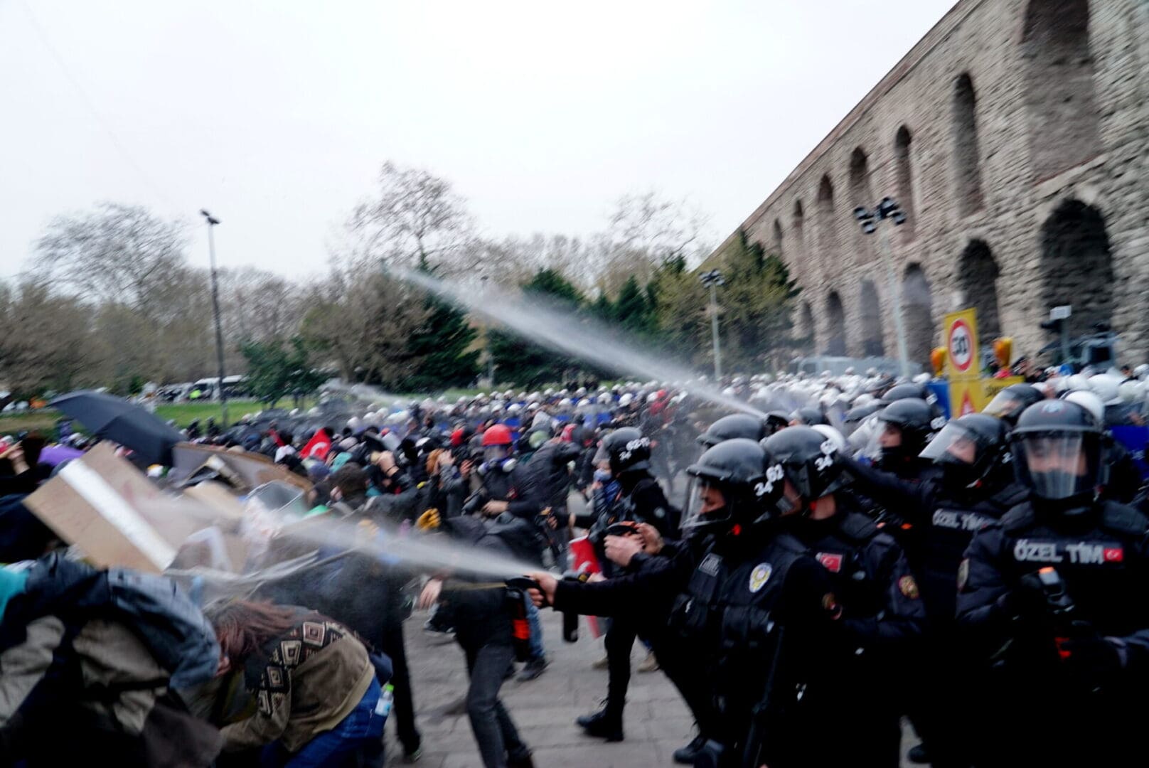 Police wearing helmets spray liquid at protesters under a gray sky.