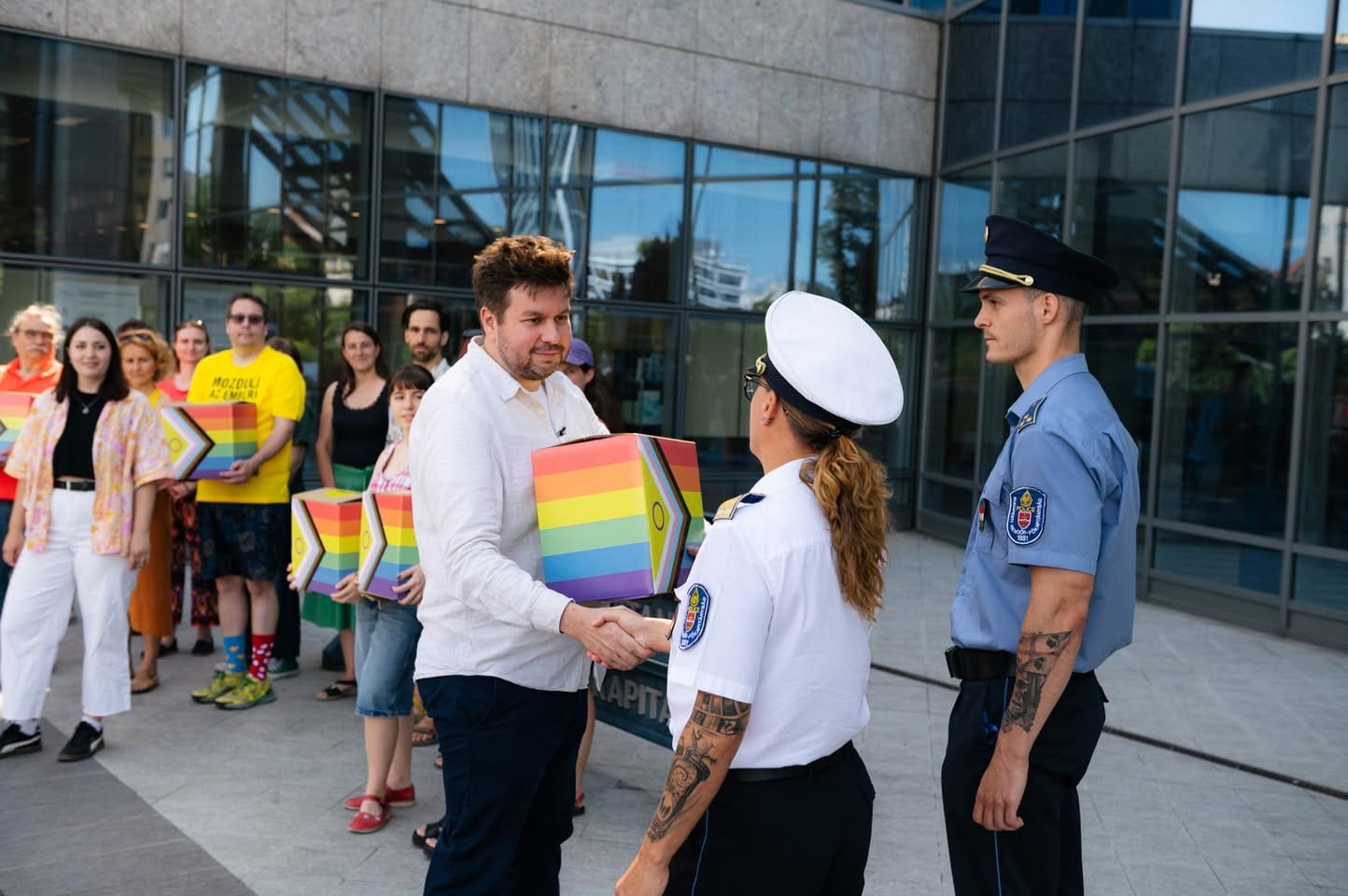 A man carrying a rainbow colored box shakes hands with a police woman in a white uniform, as more people carrying rainbow colored boxes look on, and another police officer in a blue uniform watches.