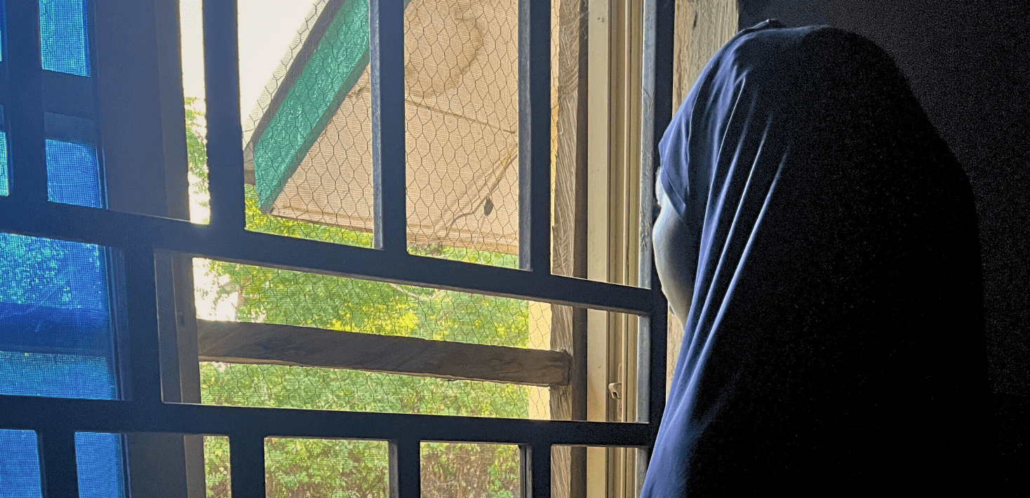 A girl in a blue headscarf in a darkened room looks through bars on a window.