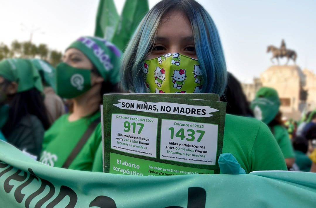 At a protest, a woman with blue dyed hair wearing a green facemask and shirt which is the color of the movement to access and legalize abortions, looks into the camera and holds up a sign with statistics about how many children 14 years old and younger have been forced to become mothers.