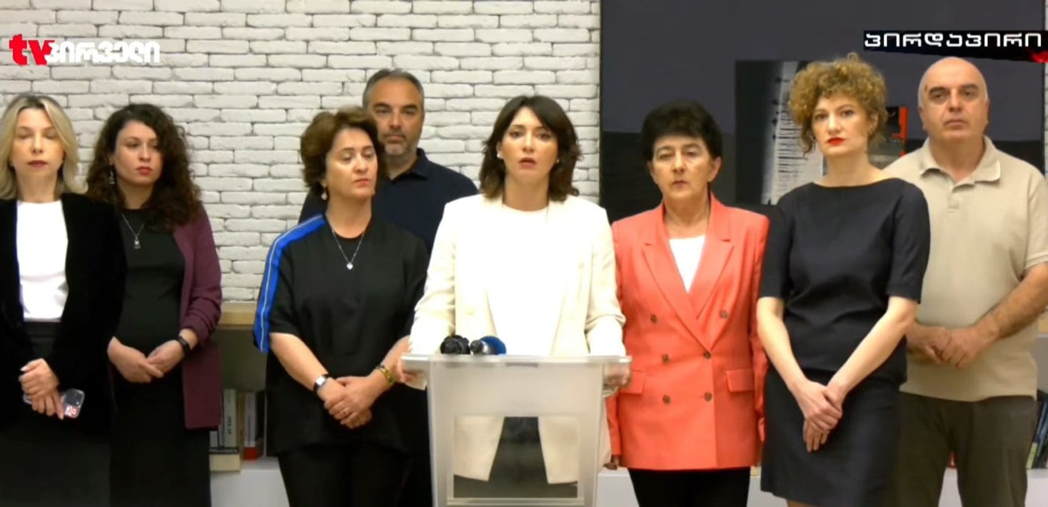 A group of eight people stands at a press conference looking serious and solemn, with a woman speaking at a podium looking directly ahead.