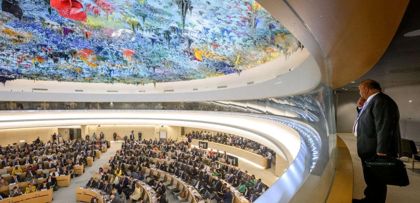 A man in a suit stands in a side gallery overlooking the The Human Rights and Alliance of Civilizations Room at the United Nations complex in Geneva, where people are seated on the floor of the hall beneath the brightly colored painted and sculpted ceiling.