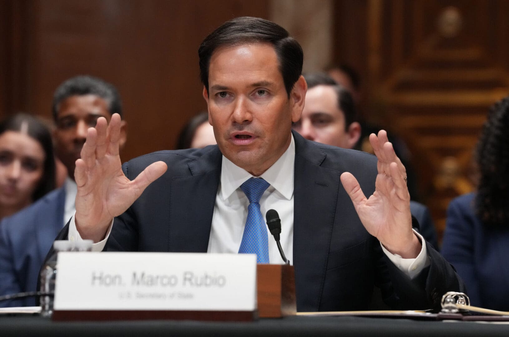 US Secretary of State Marco Rubio, wearing a blue suit and tie, sits at a table in a hearing and holds his hands up while answering questions into a microphone, with a group of onlookers seated behind him.