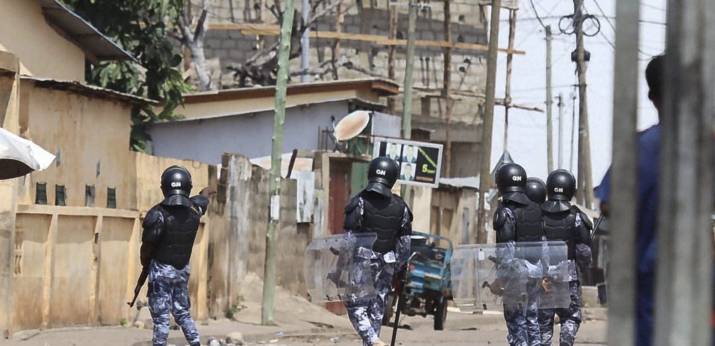 A group of police officers wearing helmets and protective gear, one carrying a rifle and the others carrying shields, patrol a deserted street amid telephone poles, concrete buildings, and a construction site. One person stands among telephone poles watching from the shadows.