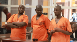 Three men stand in a courtroom wearing orange prison uniforms, one of them has his arms crossed, the one in the middle has his hands behind his back, and the third scratches his fingers.