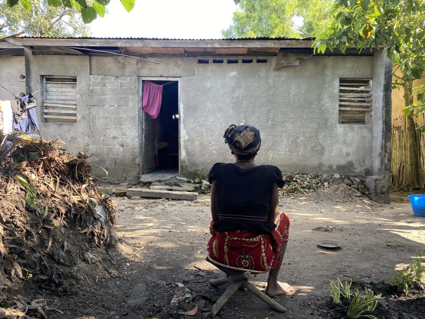 A woman in a black shirt, red wrap, and headscarf sits on a wooden stool in a dirt yard facing a small concrete house with clothes hanging on a line. There are green trees around, a bamboo fence, some plants poking up in a garden, and a pile of dirt and dry leaves in the yard.
