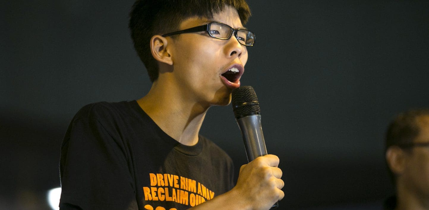 Joshua Wong, who is a young clean shaven man wearing black rectangular framed glasses and a black tshirt with orange writing, leans forward as he grips a microphone and speaks passionately into it.