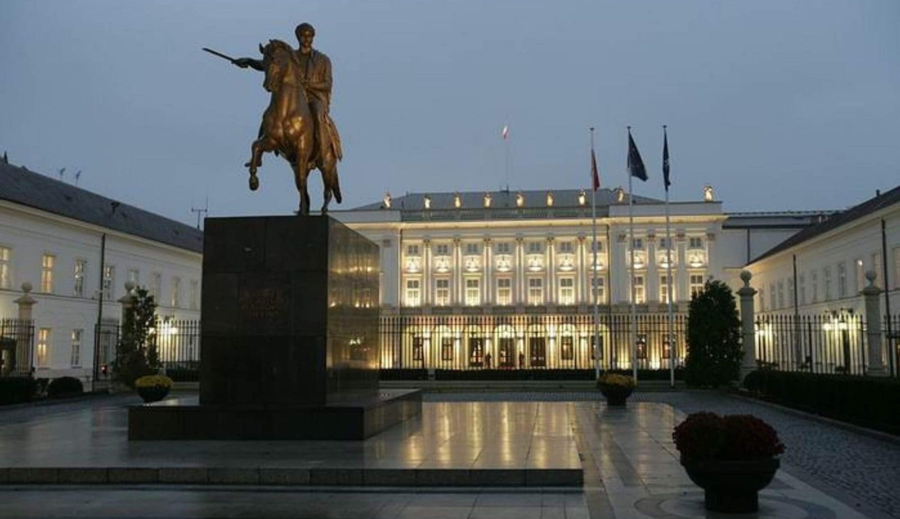 Poland's presidential palace at dusk. A large, darkened statue of a man on horseback pointing a sword forward infront of a large, stately white building with flags, lit up by lights which are reflected in a central plaza.
