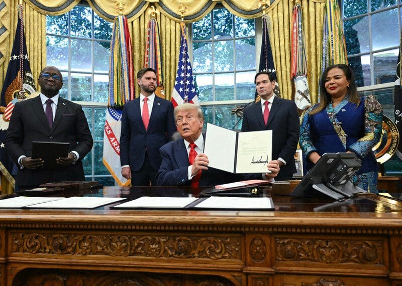 US President Donald Trump, sitting at his desk in the Oval Office, holds up a document with his signature, with US, DRC, and Rwandan officials behind him.
