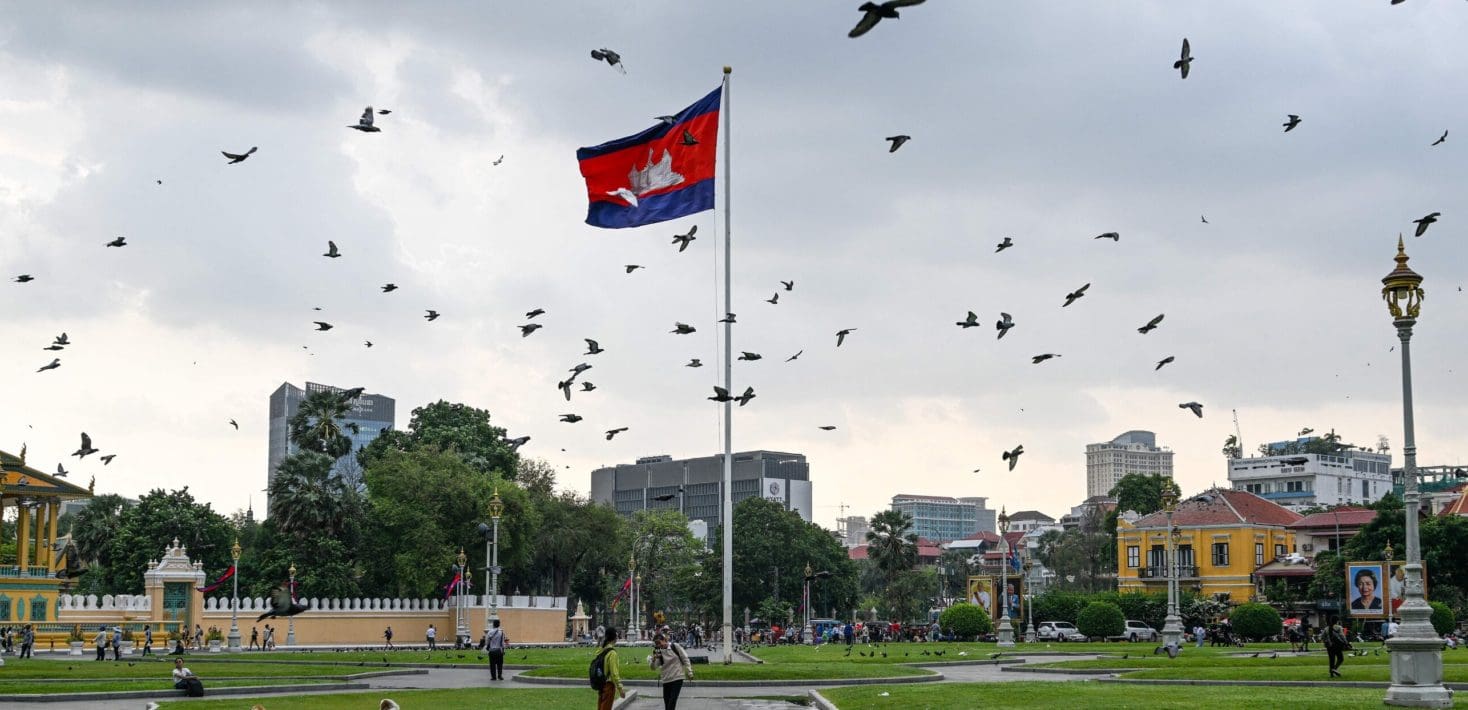 The Cambodian flag waves atop a flagpole at dawn surrounded by flying pigeons in a grassy park with buildings on the horizon.