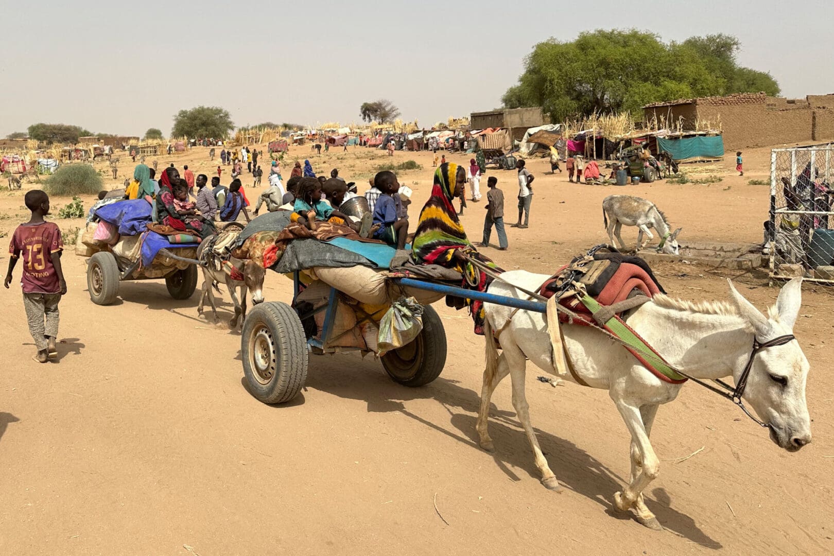 A donkey pulls a cart loaded with women and children through a sparse village of mud brick and thatch buildings.