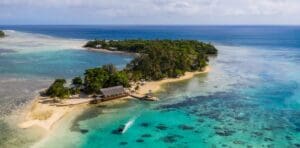 A tropical island lined with beaches and filled with green palm trees surrounded by clear turquoise waters under a blue sky with white clouds.