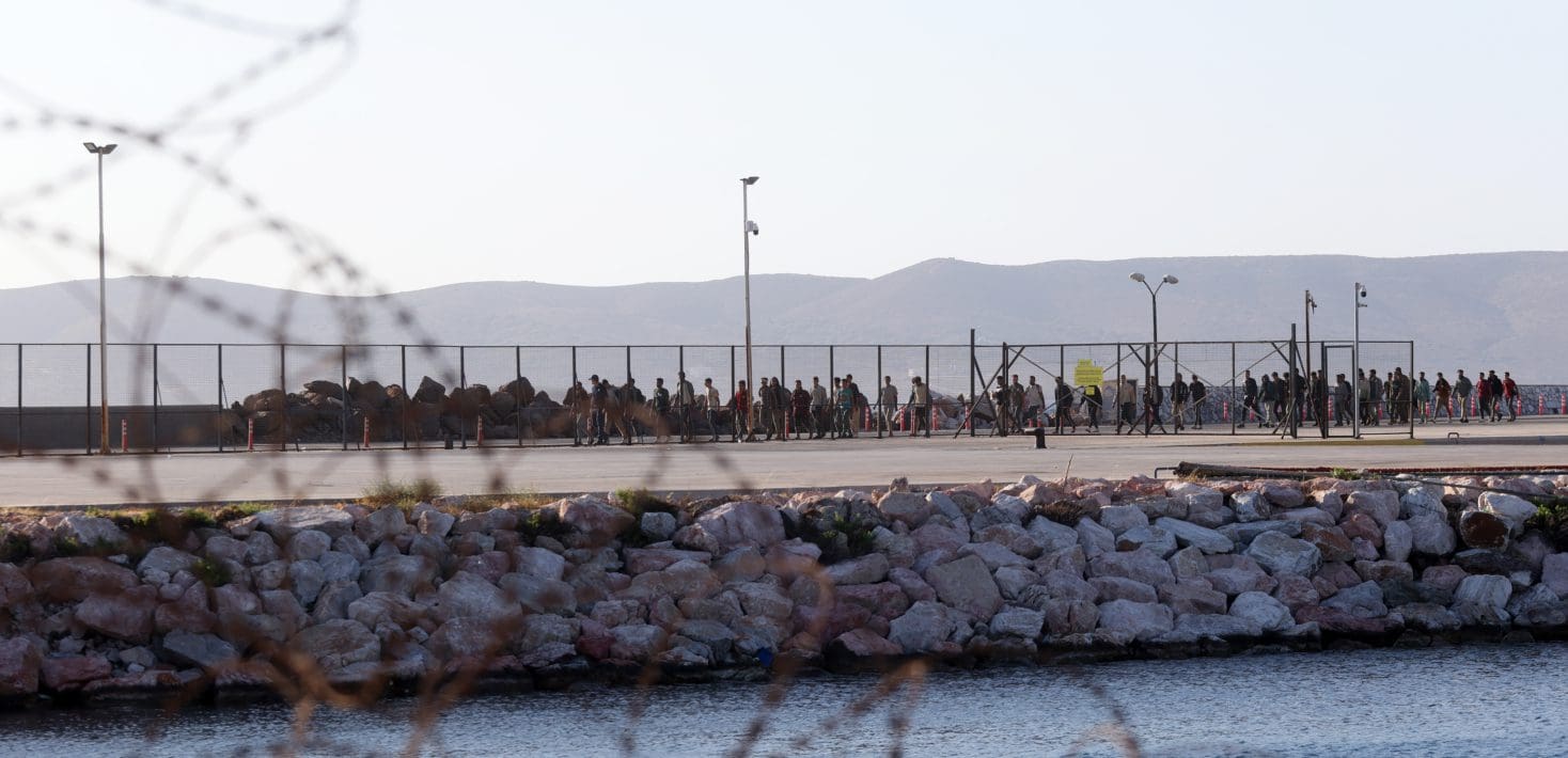 A line of people stand by a tall fence, seen through barbed wire, by the sea and in front of low mountains at dawn.