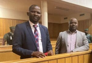 Wearing suits and ties, and looking with slight smiles into the camera, Mduduzi Maceda Mabuza and Mthandeni Dube stand in the dock in a courtroom.