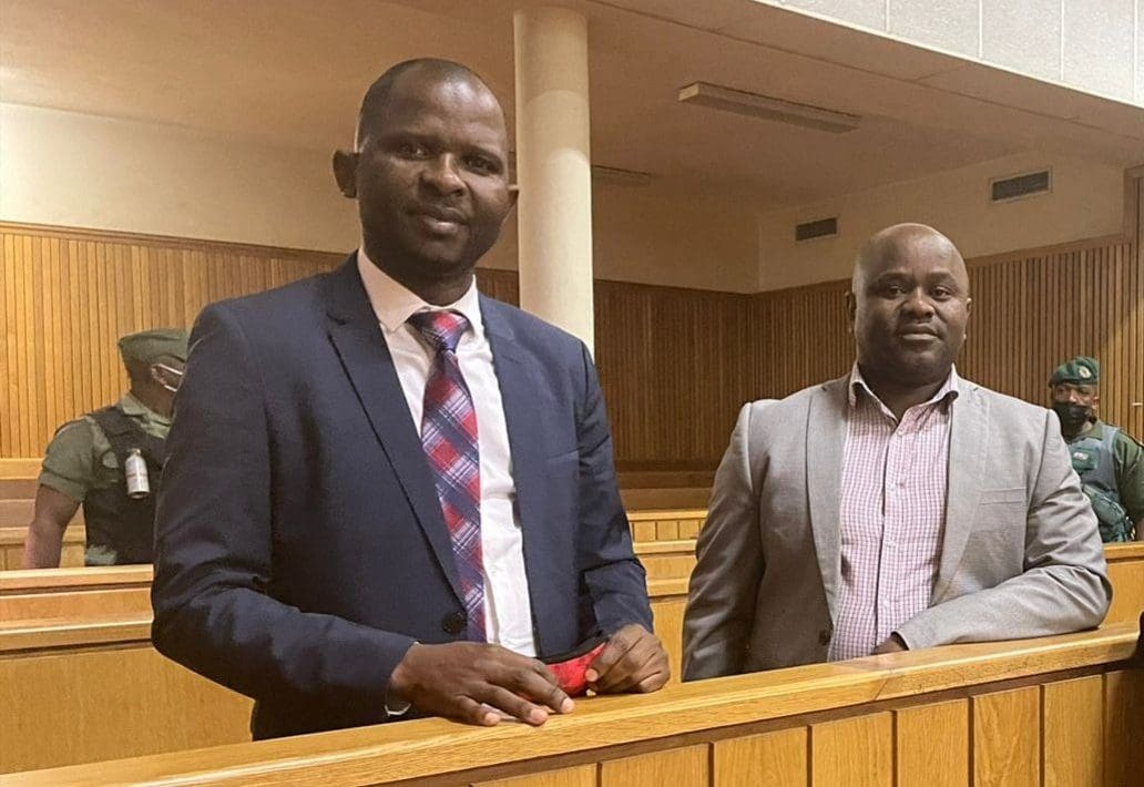 Wearing suits and ties, and looking with slight smiles into the camera, Mduduzi Maceda Mabuza and Mthandeni Dube stand in the dock in a courtroom.