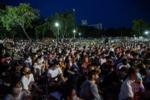 Thousands of young people sit in a park at night at a protest, wearing medical masks.