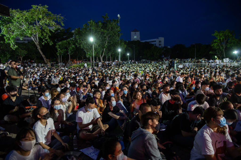 Thousands of young people sit in a park at night at a protest, wearing medical masks.