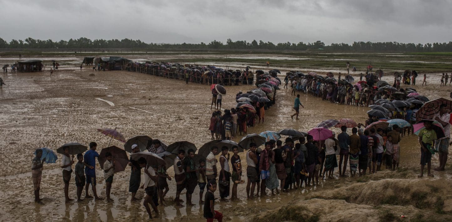 Hundreds of people stand in line under umbrellas in a wet muddy field beneath a gray sky in the rain. It is clearly windy, and the people are clutching themselves, cold.