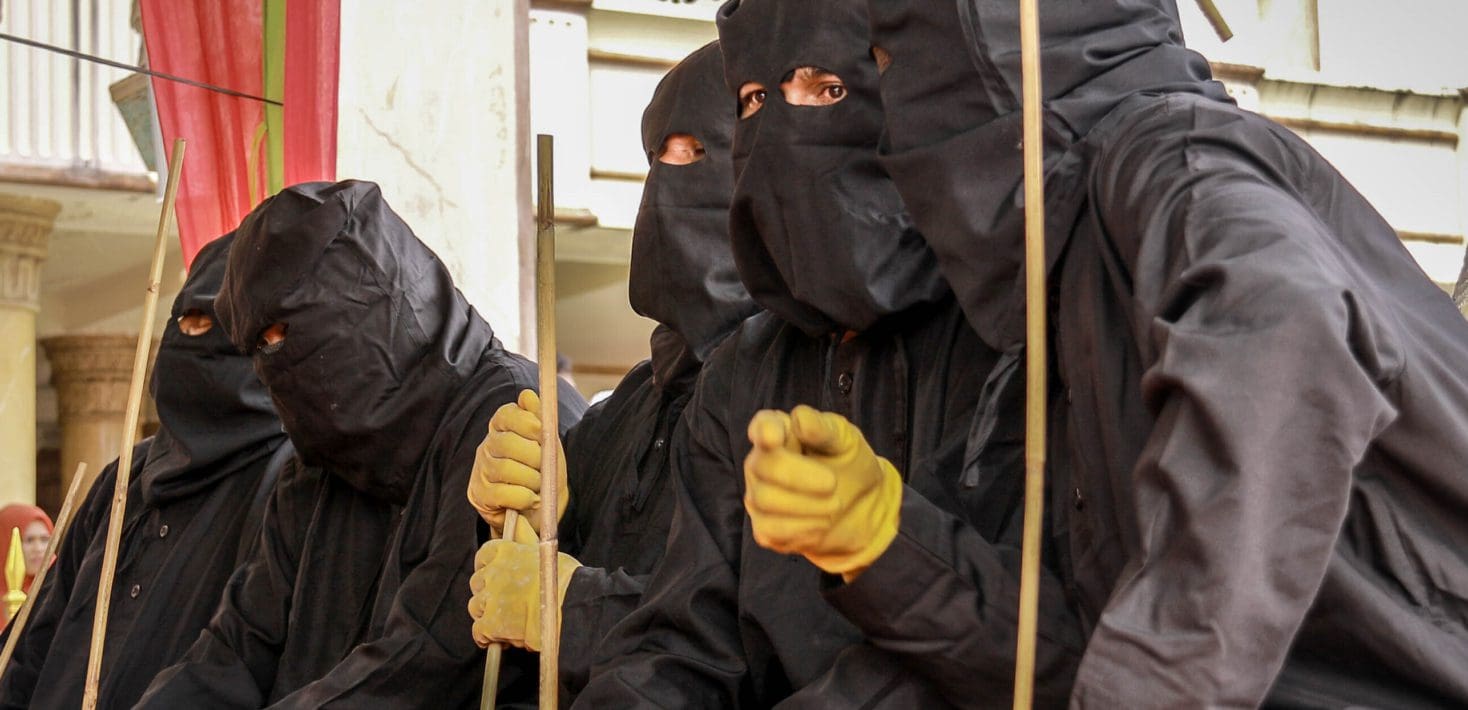 A row of men wearing black hoods and cloaks and leather gloves hold long thin bamboo canes to flog people with.