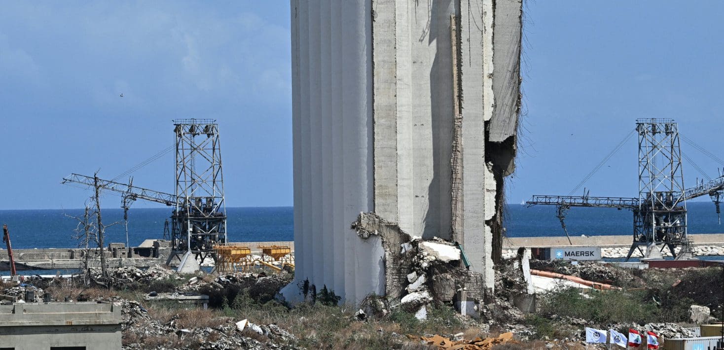 Piles of concrete and torn metal stand next to shipping container cranes with the blue sea in the background.