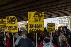 Protesters wearing keffiyahs walk below an underpass holding yellow and black signs that say 