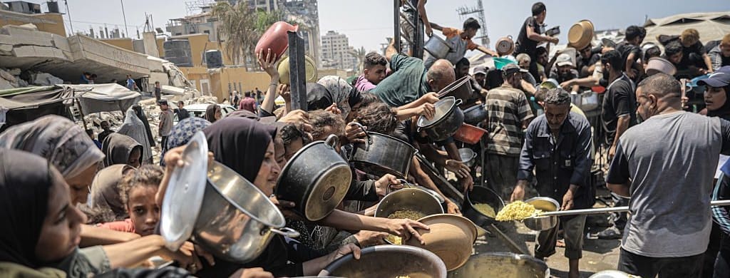 A crowd of people stretch out their arms holding pots and pans in hopes of receiving food.