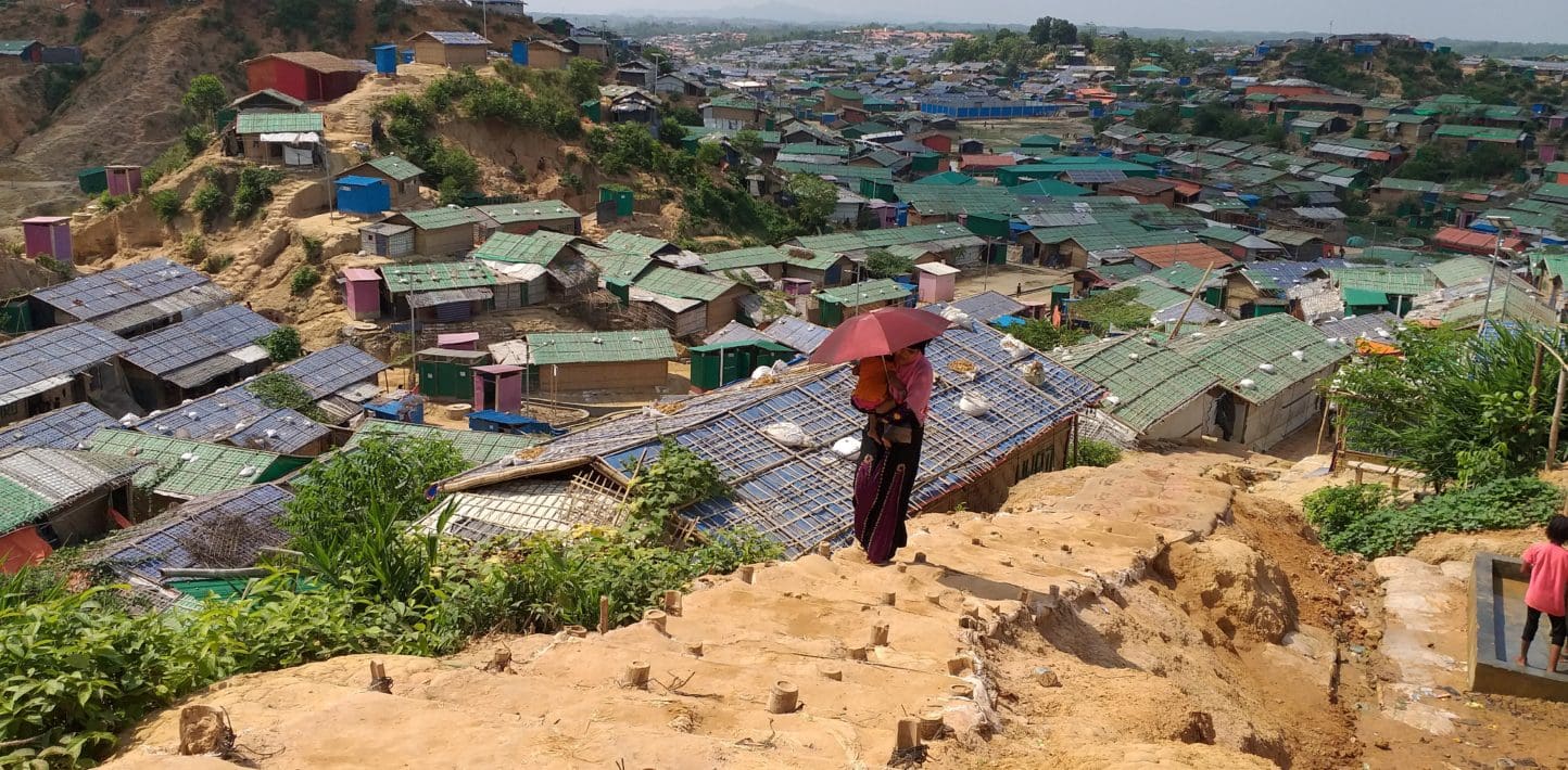 A woman carries a baby under an umbrella to shield from the bright sun as she walks up a dirt path on a steep hillside. Behind her are seemingly endless shacks and huts with plastic sheet walls and metal roofs, built onto hillsides and in small valleys, stretching to the horizon.