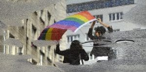 Two people waving a rainbow flag are reflected in a puddle of water on a city street.