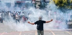 A person in a black tshirt and a colorful bucket hat stands with arms outstretched to their sides in a street filled with white tear gas smoke.