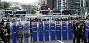 A line of police in black uniforms blocks a street with tall blue plastic shields, in front of metal barricades and rows of armored vehicles.